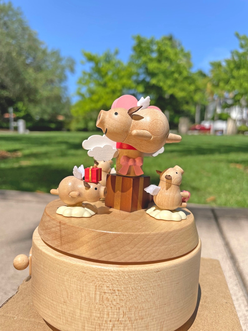 May include: A wooden music box with a whimsical design featuring three pig figurines with wings. The top pig is flying with a pink bow and a red gift box. The music box is made of light brown wood and has a round shape.