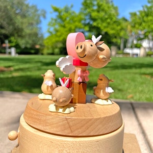 May include: A wooden music box with a pink heart and two pig figurines on top. Two birds are on either side of the base. The music box is made of light wood and has a natural finish.
