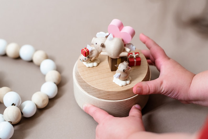 May include: A wooden music box with a pink heart and three pig figurines. The pig figurines are holding gifts and have wings. The music box is on a light brown surface.