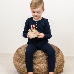 May include: A young child sits on a woven rope stool and holds a wooden musical toy with small animal figurines. The toy is painted in natural wood tones with pink and yellow accents.