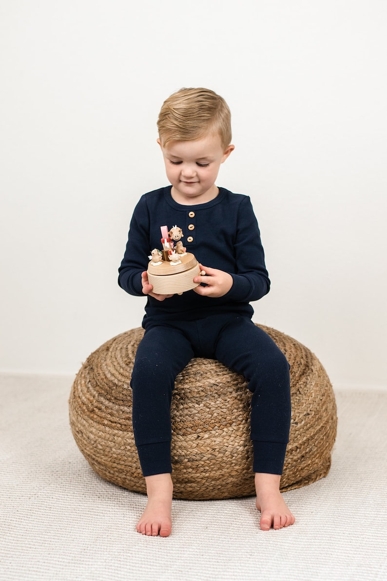 May include: A young child sits on a woven rope stool and holds a wooden musical toy with small animal figurines. The toy is painted in natural wood tones with pink and yellow accents.