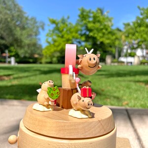 May include: A wooden music box with a rotating carousel featuring three pig figurines. The pig figurines are painted in shades of brown and pink. One pig is holding a bouquet of green leaves, another is holding a red gift box, and the third is on a pink ribbon.