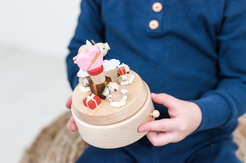 May include: A wooden music box with a pink heart and a cupid's arrow on top. The music box is decorated with small wooden animals and clouds. There are also small presents on the music box.