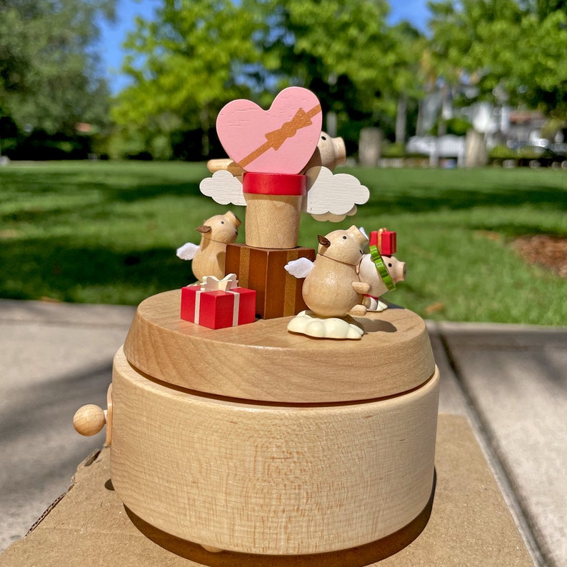 May include: A wooden music box with a pink heart and three pig figurines. The pigs are wearing angel wings and holding gifts. The music box is on a brown surface.