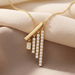 Peut inclure: Un collier doré composé d'une barre horizontale à laquelle sont suspendues quatre barres verticales de longueurs différentes. Trois de ces barres verticales sont incrustées de petites pierres claires, tandis que la plus courte porte une pierre taille baguette.
