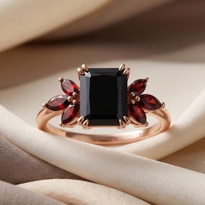 Könnte beinhalten: Ein Ring aus Roségold mit einem großen, rechteckigen schwarzen Mittelstein, der auf jeder Seite von drei tiefroten Steinen im Marquise-Schliff flankiert wird. Die Ringschiene ist schmal und poliert, die Steine sind in Krappen gefasst.