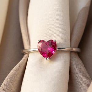 Peut inclure: Une bague en argent ornée d'une pierre précieuse rouge vif en forme de cœur, fixée par des griffes. L'anneau est lisse et poli, présentant une finition métallique nette qui complète la pierre en forme de cœur rouge profond et facettée.