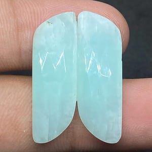 Peut inclure: Deux pierres précieuses polies, de couleur bleu clair, avec un dessus incurvé et une base plate. Elles sont tenues par des doigts. Les pierres présentent un motif interne subtil et une surface lisse et réfléchissante. La forme rappelle un papillon.