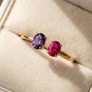 May include: A gold open-style ring featuring two oval-cut gemstones, one purple amethyst and one vibrant pink ruby, each secured in a four-prong setting. The ring is displayed inside a cream-colored velvet jewelry box with a soft, padded interior.