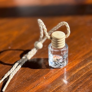 Op de afbeelding: Een klein glazen flesje met een houten deksel en een touwtje eraan. Het flesje is gevuld met een heldere vloeistof en kleine, kleurrijke voorwerpen.