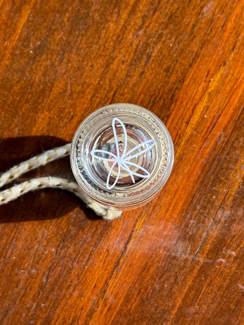 May include: A clear glass bottle stopper with a white floral design. The stopper has a rope attached to it.