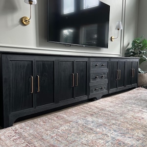 May include: A long black cabinet with multiple doors and drawers, featuring gold hardware. The cabinet is positioned in front of a rug with a multi-colored pattern.