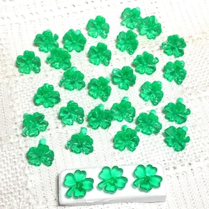 May include: A collection of translucent green clover-shaped beads. The beads are scattered across a white surface, with a row of three clovers attached to a white base. The beads have a glossy finish.