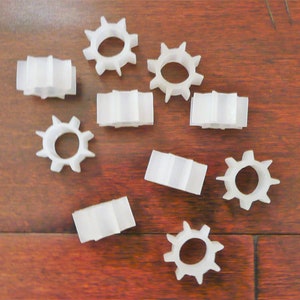 May include: A collection of ten white plastic gears of various sizes and shapes, arranged on a brown wooden surface.