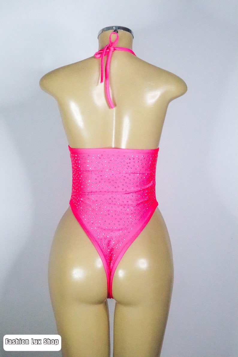 May include: A hot pink bodysuit with a halter neck and high-cut leg. The garment is embellished with small, sparkling rhinestones. The mannequin is displayed against a plain white background. The text "Fashion Lux Shop" is visible at the bottom.