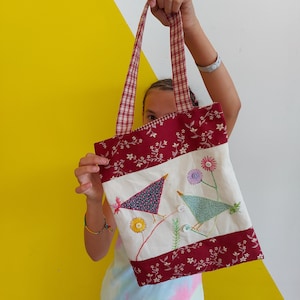 Puede incluir: Un bolso tote hecho a mano con un panel central crema con p&aacute;jaros bordados y detalles florales. Los bordes superior e inferior son de color rojo oscuro con un estampado floral, y las asas son a cuadros rojos y blancos.