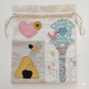 Può includere: Una borsa a coulisse color crema con un design patchwork. Il design include un cuore rosa con un girasole, un'ape, un alveare e una casetta per uccelli con un uccello. La borsa ha un cordoncino beige.