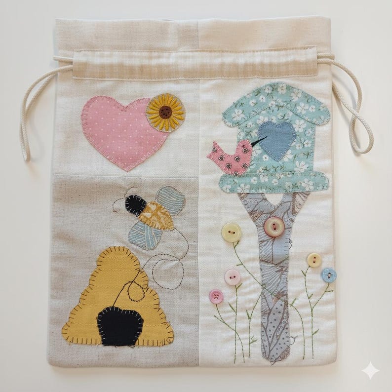 May include: A cream drawstring bag with a patchwork design. The design features a pink heart with a sunflower, a bee, a beehive, and a birdhouse with a bird. The bag has a beige drawstring.
