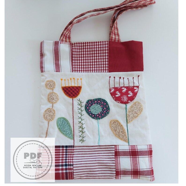 Puede incluir: Bolso tote patchwork con dise&ntilde;o floral. El bolso presenta un fondo blanco con flores bordadas en rojo, azul y beige. Las asas y el borde superior son de tela a cuadros rojos y blancos. El bolso tiene un logotipo PDF.
