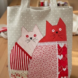 Peut inclure: Sac fourre-tout beige à pois blancs avec deux motifs de chats appliqués. Un chat est rouge à pois blancs et l'autre est rouge et blanc rayé. Les chats ont des yeux en bouton et des moustaches brodées.