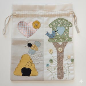 May include: A cream drawstring bag with patchwork designs. The bag features a heart, a sunflower, a bee, and a birdhouse with a bird. The designs are made of various fabrics and embellished with buttons and embroidery.