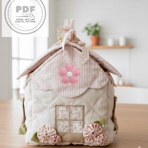 Può includere: Una teiera in tessuto trapuntato a forma di casa, in beige con un'applicazione floreale rosa. Il tetto è a righe con un bordo smerlato. La teiera ha una finestra e abbellimenti floreali, con il testo "MOIRA SEW LAB DIGITAL PATTERN".