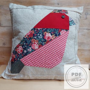 Könnte beinhalten: Dekoratives quadratisches Kissen mit einem Patchwork-Vogeldesign. Der Vogel hat einen roten Filzkopf, einen Körper mit Blumenmuster und einen rot-weiß karierten Flügel. Das Kissen ist beige mit einem dezenten Punktmuster und gesteppten Details.