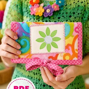 May include: A colorful, handmade wallet with a floral design. The wallet features a central square with a green embroidered flower on a cream background, surrounded by vibrant patterns. A pink polka-dot bow adds a decorative touch. The image also includes a floral necklace.
