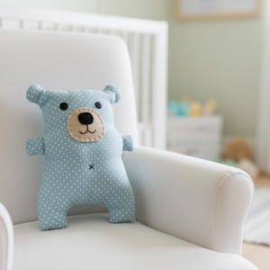 Puede incluir: Un peluche con forma de oso azul claro con lunares blancos, ojos negros y hocico beige. El peluche está sobre un sillón blanco en un cuarto de niños. El oso tiene brazos y piernas pequeños y una cruz negra cosida.