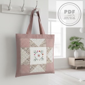 Puede incluir: Un bolso tote rosa empolvado con un diseño de patchwork. El cuadrado central presenta un diseño floral bordado. El bolso está colgado en una pared blanca con un perchero de madera. El bolso tiene un patrón digital PDF.