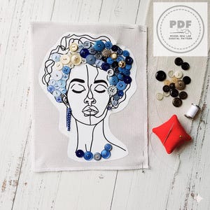 Può includere: Un'opera d'arte a tecnica mista con un disegno a tratto del volto di una donna adornato con bottoni blu e bianchi. I bottoni formano i capelli e la collana. Sono presenti anche un puntaspilli rosso, filo e bottoni aggiuntivi.