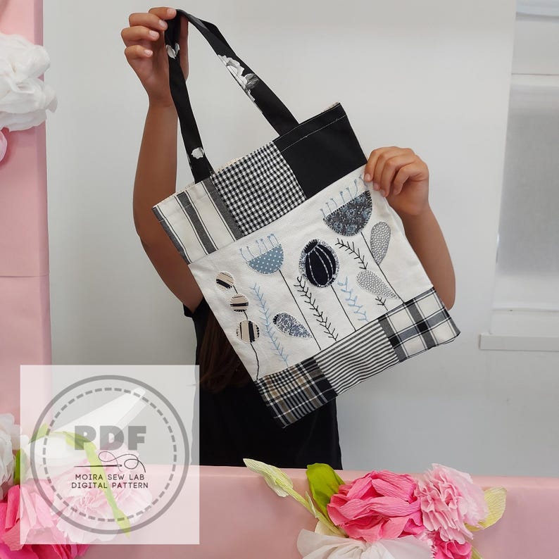 Può includere: Una borsa tote patchwork con un design floreale. La borsa presenta una varietà di tessuti, tra cui quadretti bianchi e neri, righe e motivi floreali. I manici sono neri con un motivo floreale bianco. La borsa è tenuta da una persona.