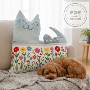 Può includere: Un cuscino a forma di gatto blu chiaro con tratti ricamati e una coda ornata di bottoni. Il cuscino presenta un bordo ricamato floreale con fiori rossi, gialli, blu e rosa. Un piccolo cane marrone sta dormendo su una coperta grigia.