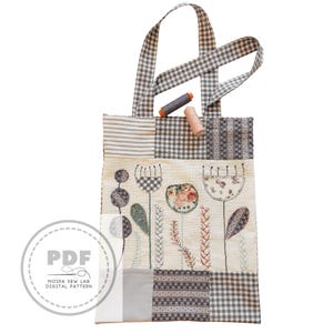Puede incluir: Un bolso tote patchwork con un dise&ntilde;o floral y asas de cuadros. El bolso presenta flores bordadas y patrones geom&eacute;tricos en tonos neutros. Dos carretes de hilo descansan en el borde superior. El bolso tiene un logotipo "PDF".