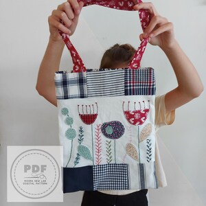 Puede incluir: Un bolso tote patchwork con bordado floral. El bolso presenta un fondo blanco con flores bordadas en rojo, azul y verde. La parte superior y las asas son de tela estampada en rojo y azul. El bolso es sostenido por una persona.