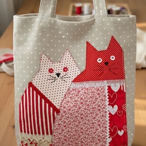 May include: A beige tote bag with a white polka dot pattern featuring an appliqué of two cats. One cat is white with red dots, and the other is solid red. The cats have button eyes, stitched whiskers, and are decorated with red striped and heart-patterned fabric.