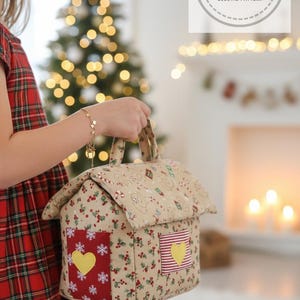 Puede incluir: Una bolsa de tela beige con forma de casa con asa, con un estampado navideño de bayas rojas, hojas verdes y copos de nieve. La bolsa tiene detalles a rayas rojas y blancas y copos de nieve con detalles de corazón amarillo. La bolsa se sostiene frente a un árbol de Navidad.