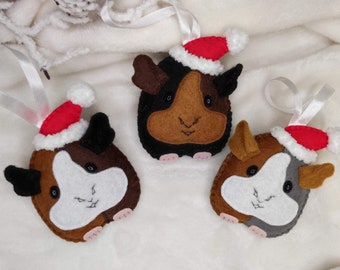 May include: Three felt guinea pig ornaments with Santa hats. The ornaments are brown, black, and white with embroidered facial features. Each ornament has a white ribbon for hanging. Festive holiday decor.