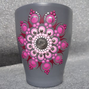 Puede incluir: Taza de cerámica gris con un diseño de mandala pintado a mano. El mandala presenta un patrón simétrico de puntos y formas rosas y plateadas, con un contorno rojo oscuro. La taza es cilíndrica y tiene una superficie lisa.