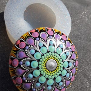 以下が含まれることがあります： 白いシリコンモールドを背景に、丸い石にカラフルなマンダラが描かれています。マンダラは、ピンク、パープル、ブルー、グリーン、イエロー、シルバーなど、さまざまな色を使用しています。マンダラは黒い縁取りで囲まれています。
