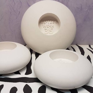 Puede incluir: Tres portavelas de cerámica blanca de diferentes tamaños. El portavelas más grande tiene el texto "GEFRÄZER WERK 35" en la parte inferior.