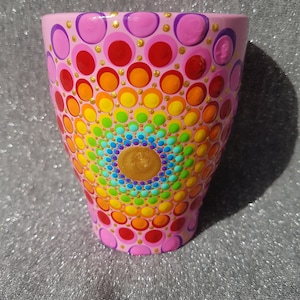 Può includere: Un vaso di ceramica rosa con un design a mandala arcobaleno dipinto sul davanti. Il mandala è composto da piccoli punti di vari colori, tra cui rosso, arancione, giallo, verde, blu, indaco e viola. Il vaso è decorato con glitter dorati.