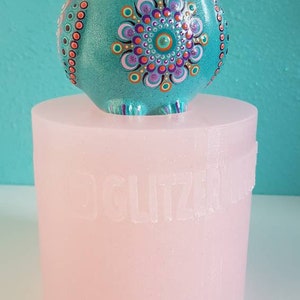 Pode incluir: Uma estatueta de coruja turquesa com um design de mandala detalhado em tons de roxo, laranja e rosa. A coruja está em cima de uma base cilíndrica rosa claro. Os olhos e as penas da coruja são intrincadamente decorados com pontos e padrões.