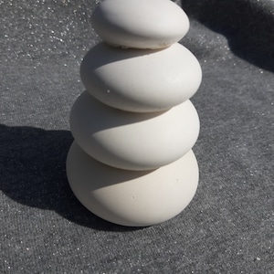 Puede incluir: Una pila de cuatro piedras redondas, lisas y blancas, dispuestas en forma de pirámide. Las piedras están sobre una superficie gris texturizada.