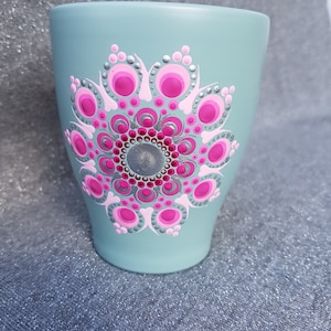 Puede incluir: Una taza de cerámica azul claro con un diseño de mandala rosa y plateado pintado en la parte delantera.