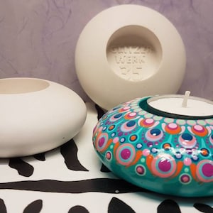 Op de afbeelding: Drie witte keramische kaarshouders, waarvan één met een teal, roze en oranje mandala-ontwerp en een witte theelichtkaars erin. De kaarshouder staat op een zwart-witte zebraprint-oppervlak.
