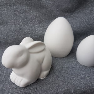 Pode incluir: Uma figurinha de coelho de cerâmica branca e dois ovos de cerâmica branca. O coelho está sentado em uma superfície cinza texturizada.