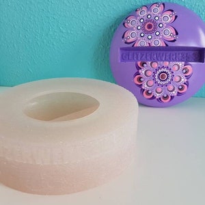 Puede incluir: Una caja redonda de color púrpura con un diseño de mandala y el texto "Glitterworks" en el lateral. La caja está abierta y el interior es un anillo blanco con un anillo más pequeño en el centro.