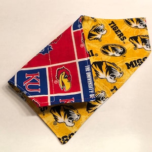May include: A red, blue, and white fabric bandana with the text "KU" and a bird logo. The bandana is folded over a yellow fabric bandana with the text "TIGERS" and a tiger logo. The University of Missouri logo is also visible.