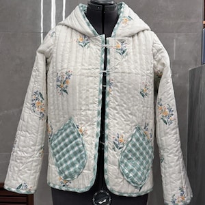 K&ouml;nnte beinhalten: Eine wei&szlig;e gesteppte Jacke mit Blumendruck und gr&uuml;nen Karotassen. Die Jacke hat eine Kapuze und einen Knopfverschluss.